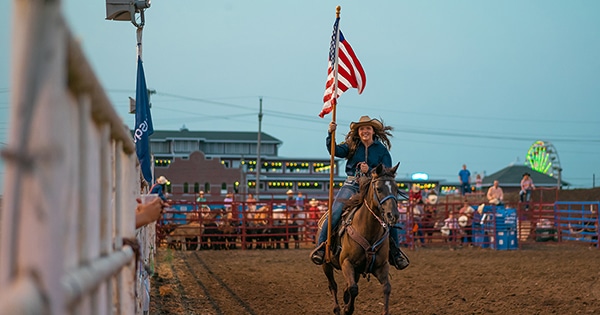 A smiling rodeo queen riding a horse and carrying the United States flag
